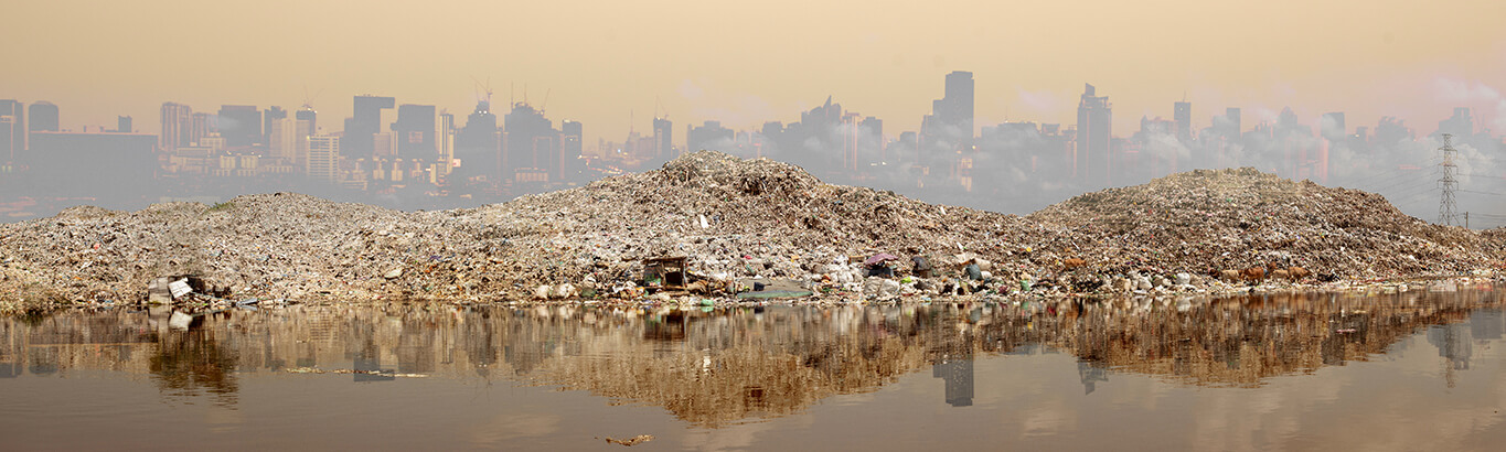 A riverbank near a city is polluted with large piles of washed-up waste.